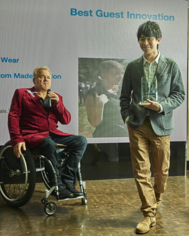 white man with blond hair and red jacket, next to Asian man in grey jacket& brown trousers